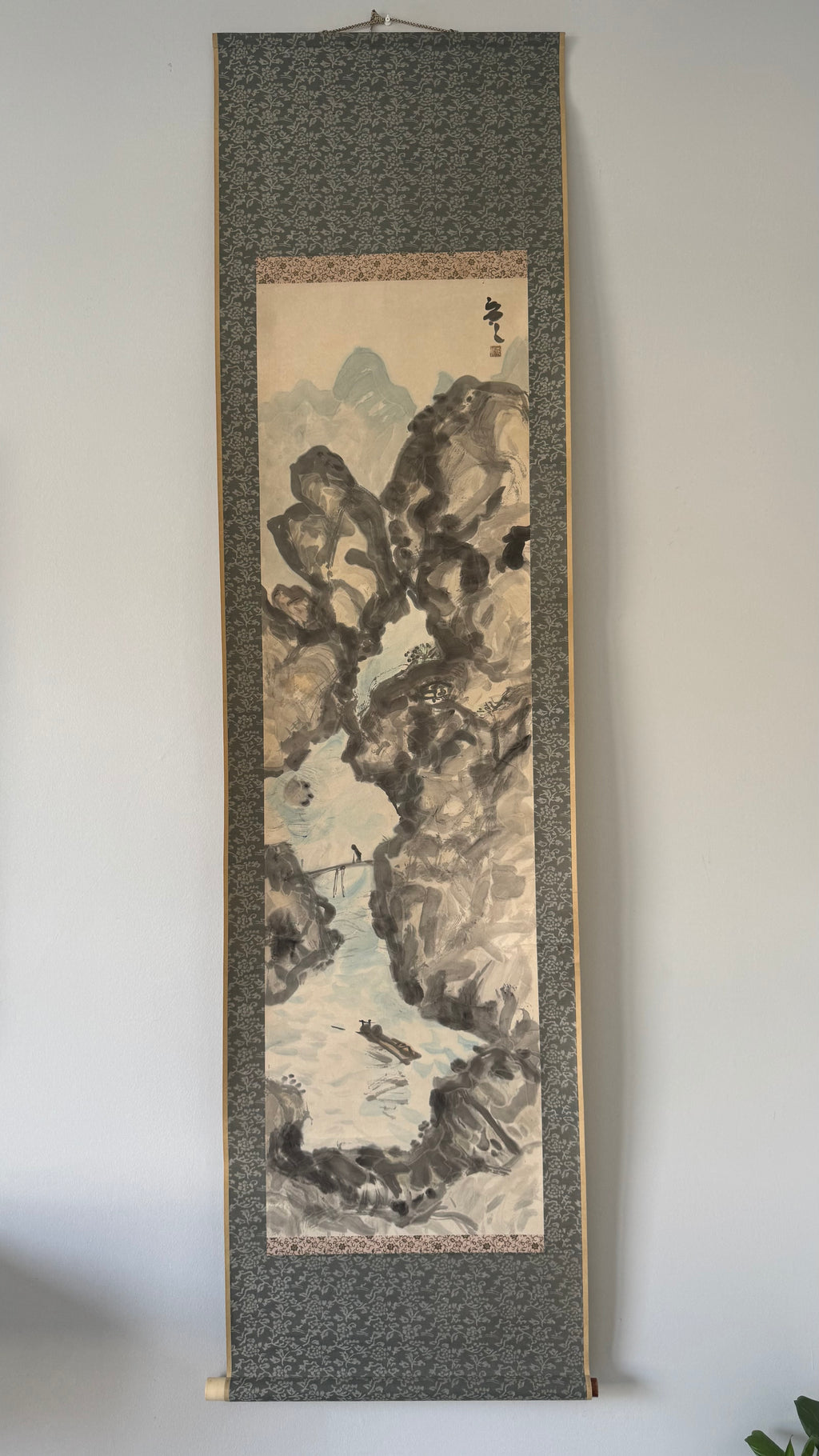 Traditional Japanes landscape painting on a vertical scroll, displayed against a plain wall.