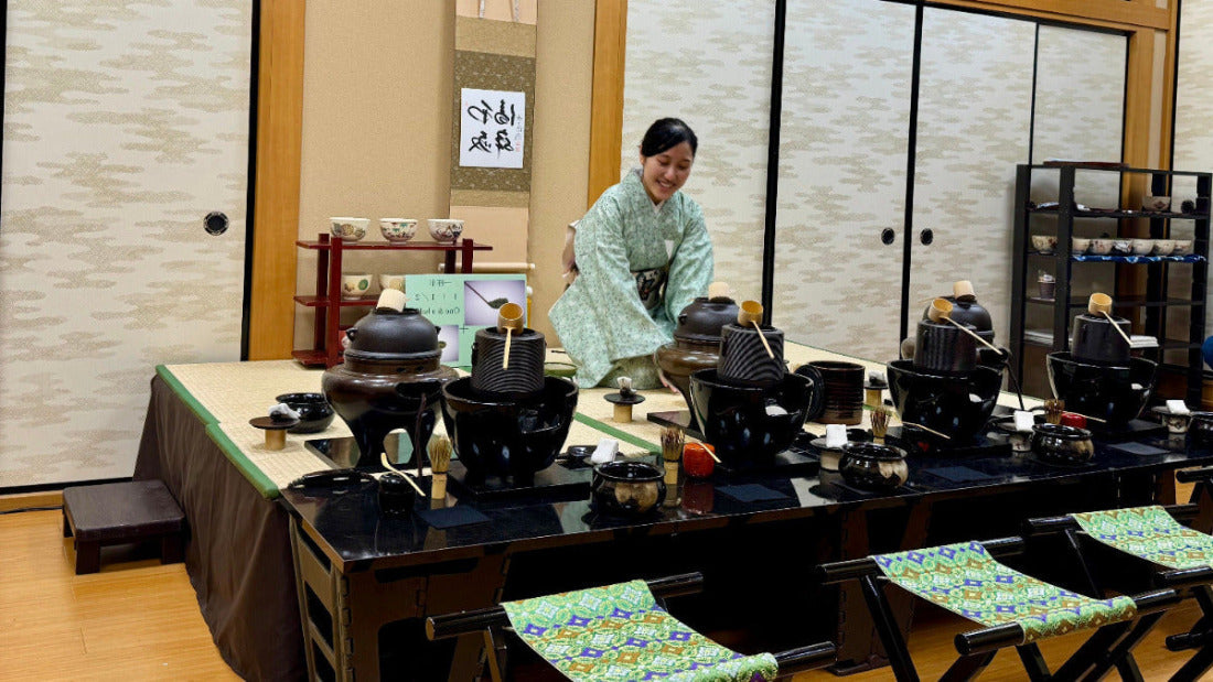 Person in traditional attire preparing tea in a room with tea-making equipment and chairs.