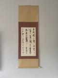 A traditional Japanese hanging scroll with gold-toned paper borders, a brown center panel, and black calligraphy on a white paper sheet, mounted against a plain light gray wall.