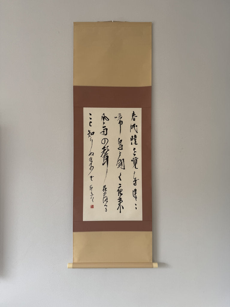 A traditional Japanese hanging scroll with gold-toned paper borders, a brown center panel, and black calligraphy on a white paper sheet, mounted against a plain light gray wall.