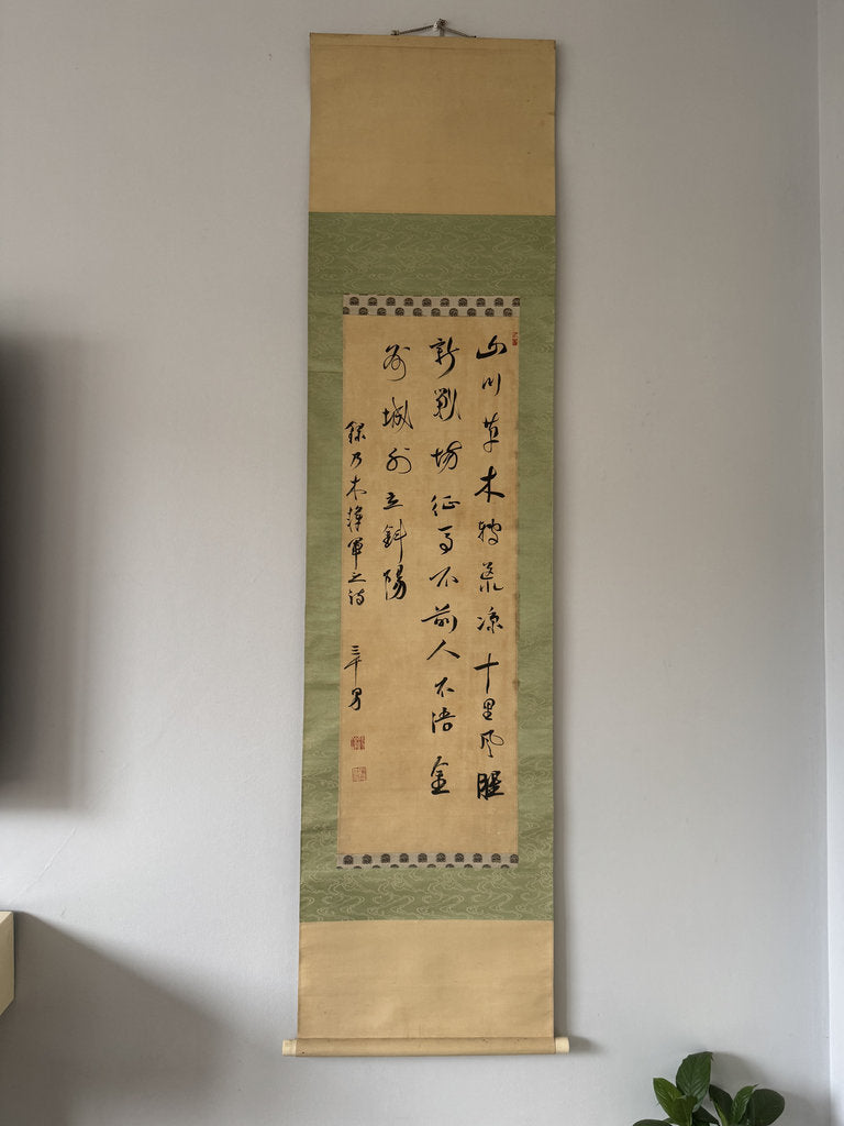 A traditional Japanese hanging scroll with black calligraphy on aged beige paper, bordered by green fabric bands, displayed on a light gray wall.