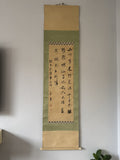 A traditional Japanese hanging scroll with black calligraphy on aged beige paper, bordered by green fabric bands, displayed on a light gray wall.