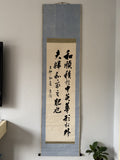 A vertical Japanese calligraphy scroll with black ink characters on aged beige paper, mounted on a textured blue-gray fabric background, hanging on a light gray wall.