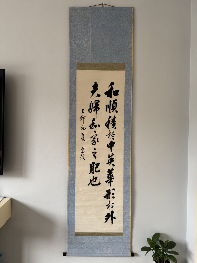 A vertical Japanese calligraphy scroll with black ink characters on aged beige paper, mounted on a textured blue-gray fabric background, hanging on a light gray wall.