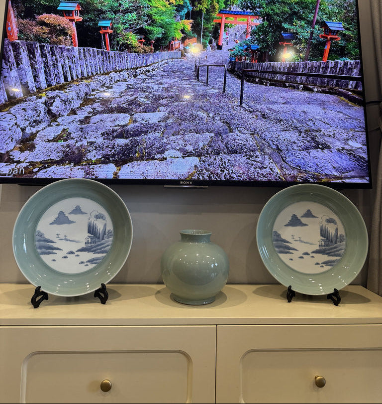 Two plates and a vase on a cabinet with a television displaying a scenic landscape in the background.