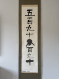 A vertical Japanese calligraphy scroll with black brush-stroke characters on an off-white paper center panel, mounted on a dark patterned fabric border against a plain light gray wall.