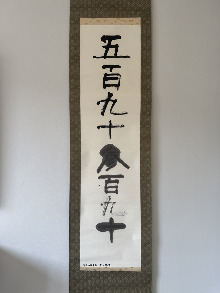 A vertical Japanese calligraphy scroll with black brush-stroke characters on an off-white paper center panel, mounted on a dark patterned fabric border against a plain light gray wall.