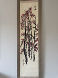 A traditional Japanese hanging scroll painting depicting red blossoms on dark branches, mounted on a light wall.