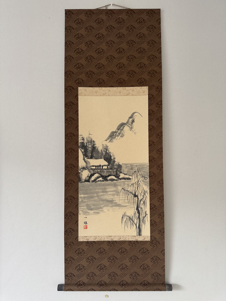 A traditional Japanese hanging scroll (kakemono) featuring a landscape painting of a house by the water, displayed against a plain white wall.