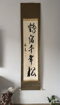 A vertical Japan calligraphy scroll hanging on a light gray wall in a home interior setting, featuring black ink characters on a beige paper background with a textured brown border.