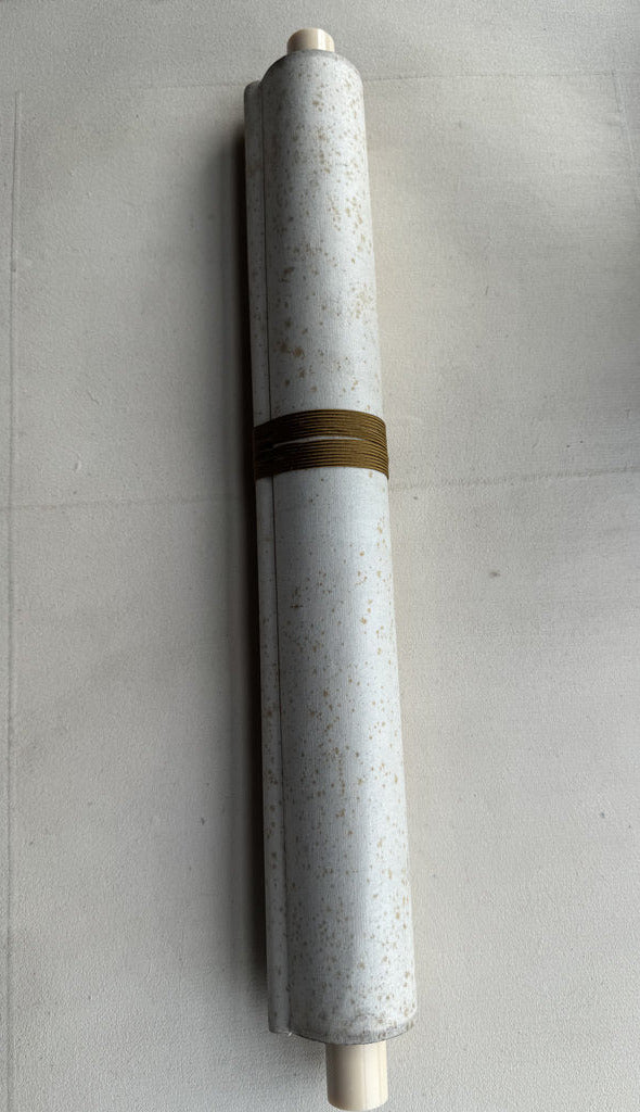 A rolled-up white fabric or wallpaper sample with small beige speckles and a metallic gold band around the middle, photographed on a light gray surface.