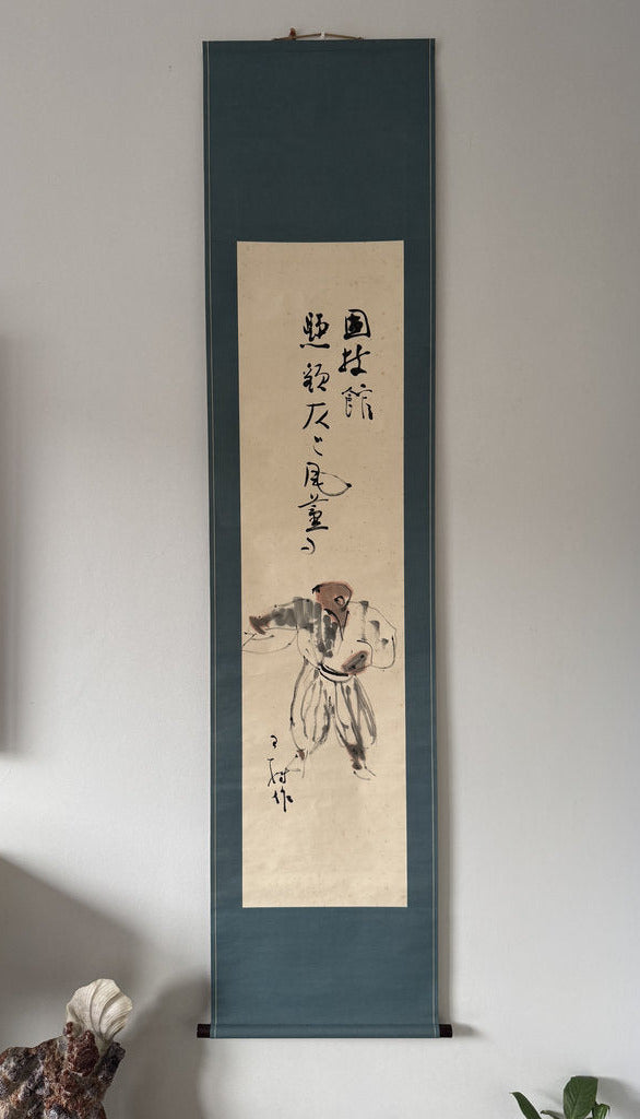 A traditional Japanes hanging scroll artwork displayed on a light gray wall. The scroll has a dark teal-blue border and a beige center panel featuring a black ink sketch of a person and vertical calligraphy.