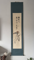 A traditional Japanes hanging scroll artwork displayed on a light gray wall. The scroll has a dark teal-blue border and a beige center panel featuring a black ink sketch of a person and vertical calligraphy.