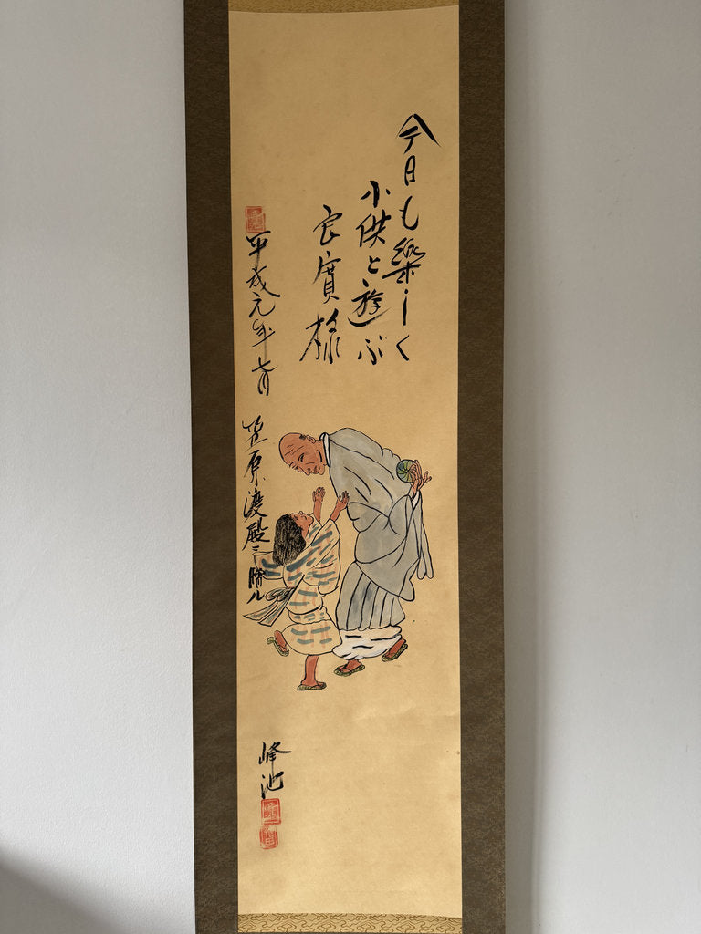 A traditional Japanese hanging scroll (kakejiku) featuring ink calligraphy and a hand-painted illustration of an elderly man and child, mounted on a light gray wall.