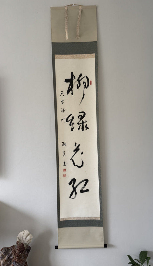 A vertical Japanese calligraphy scroll with black ink calligraphy on a beige and gray fabric background, hanging on a light gray wall in an indoor setting.