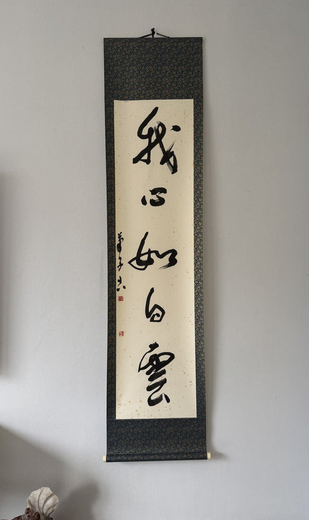 A vertical Japanese calligraphy scroll hanging on a light gray wall. The scroll has a dark textured border and a cream center panel with black calligraphy characters.