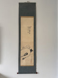 A traditional Japanese hanging scroll (kakemono) featuring a black ink sketch of a reclining figure on a beige paper background, mounted on a dark gray fabric border.