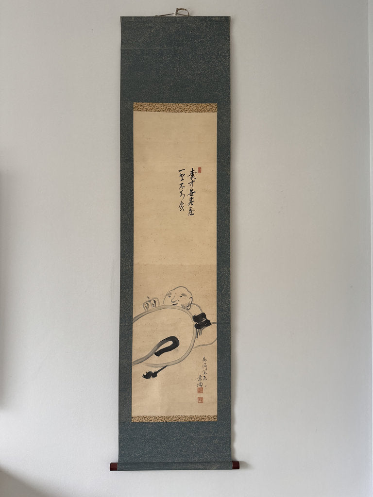 A traditional Japanese hanging scroll (kakemono) featuring a black ink sketch of a reclining figure on a beige paper background, mounted on a dark gray fabric border.