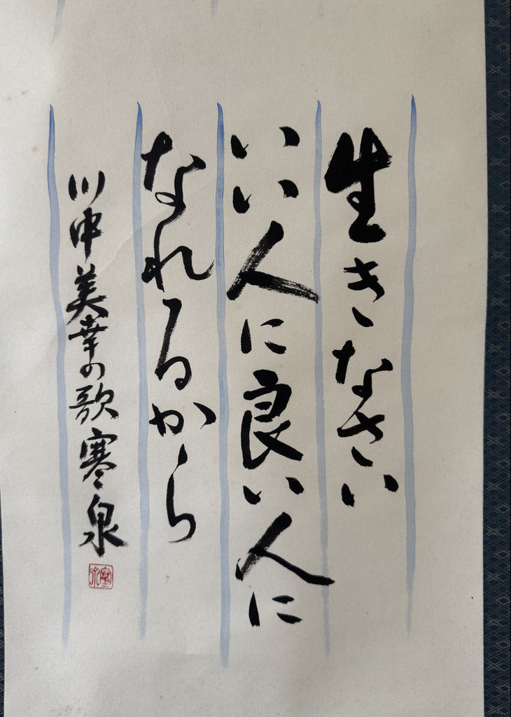 Japanese calligraphy artwork on white paper, featuring black brushstrokes, light blue vertical guide lines, and a red artist seal at the lower left, photographed against a dark patterned fabric background.
