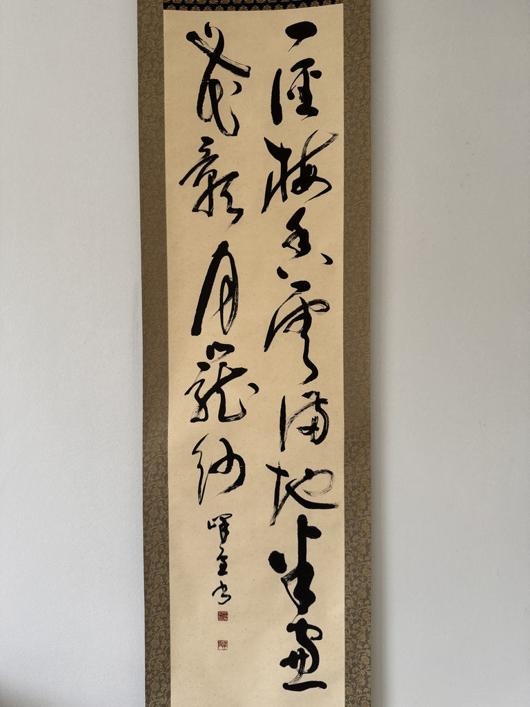 A vertical Japanese calligraphy scroll with black brushstrokes on aged beige paper, mounted on a textured brown and gray border, hanging on a plain light gray wall.