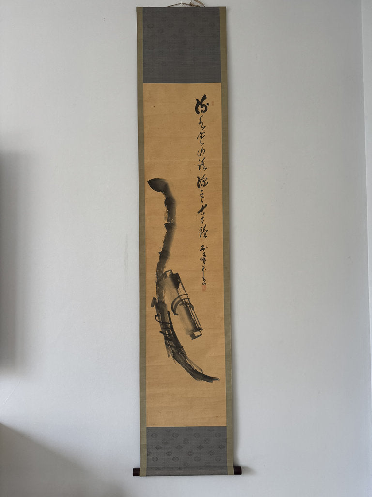 A traditional Japanse hanging scroll artwork displayed on a plain white wall. The scroll features a beige paper center with black ink brush calligraphy and a black ink painting of a curved branch or tool, framed by dark gray textured bands at the top and bottom.