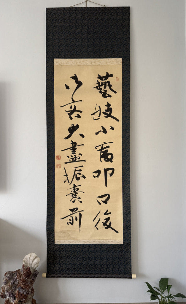 A traditional Japanese calligraphy scroll with black brushstrokes on beige paper, mounted on a dark patterned black fabric background, hanging on a light gray wall in a home interior.