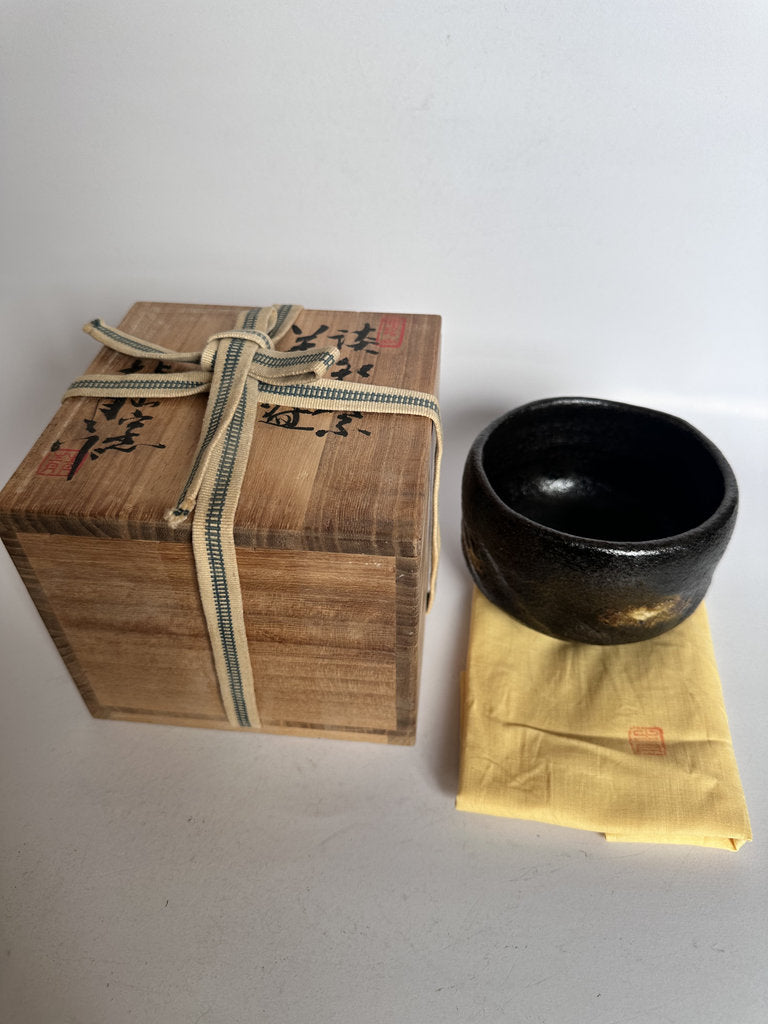 Black ceramic bowl on a yellow cloth next to a wooden box with Japanese text