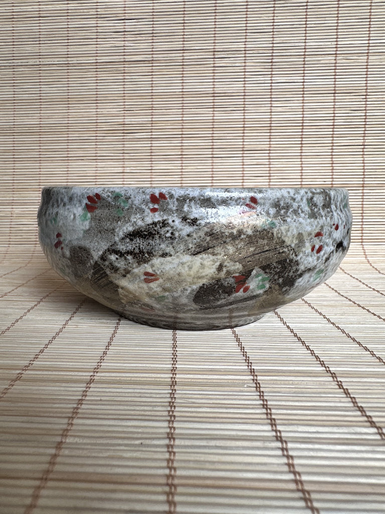 A handmade ceramic bowl with a textured, mottled glaze in gray, beige, and white, decorated with small red and green floral accents, resting on a bamboo mat.