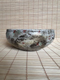 A handmade ceramic bowl with a textured, mottled glaze in gray, beige, and white, decorated with small red and green floral accents, resting on a bamboo mat.