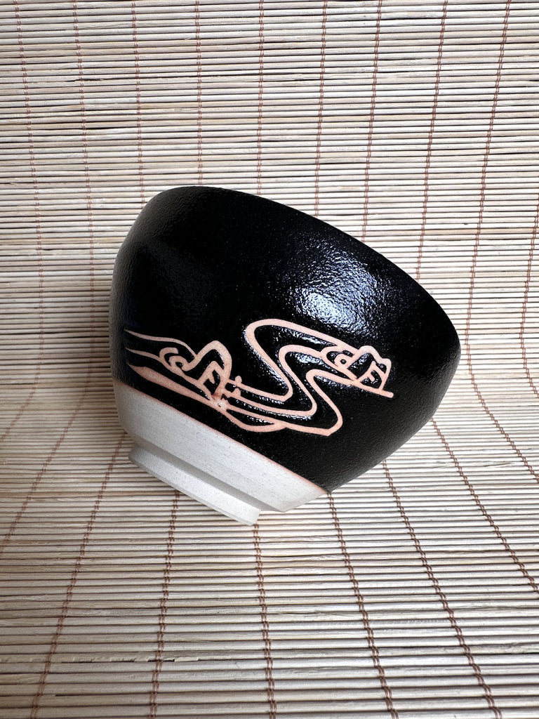 A black ceramic bowl with a white base and a pinkish-gold abstract line design, placed on a light beige bamboo mat.