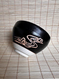 A black ceramic bowl with a white base and a pinkish-gold abstract line design, placed on a light beige bamboo mat.