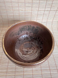A handmade ceramic bowl with a rustic, earthy brown glaze and dark speckled patterns inside, photographed from a top-down perspective on a light beige bamboo mat.
