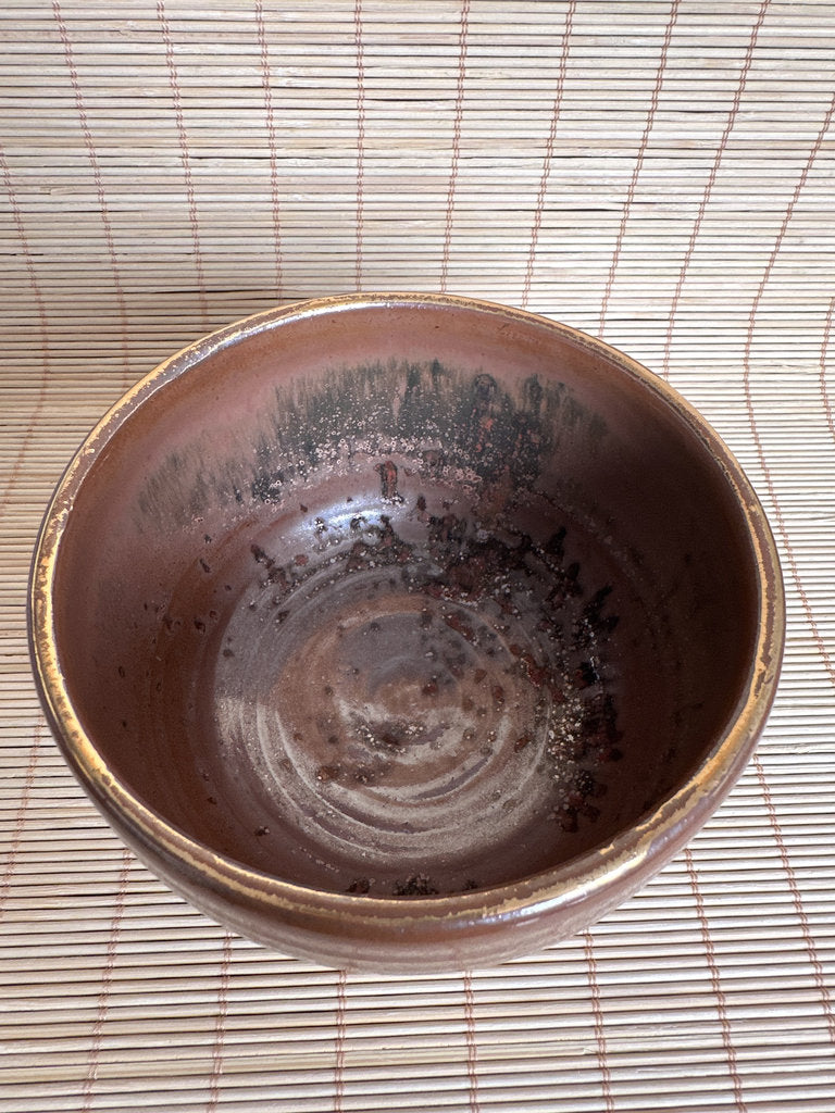 A handmade ceramic bowl with a rustic, earthy brown glaze and dark speckled patterns inside, photographed from a top-down perspective on a light beige bamboo mat.