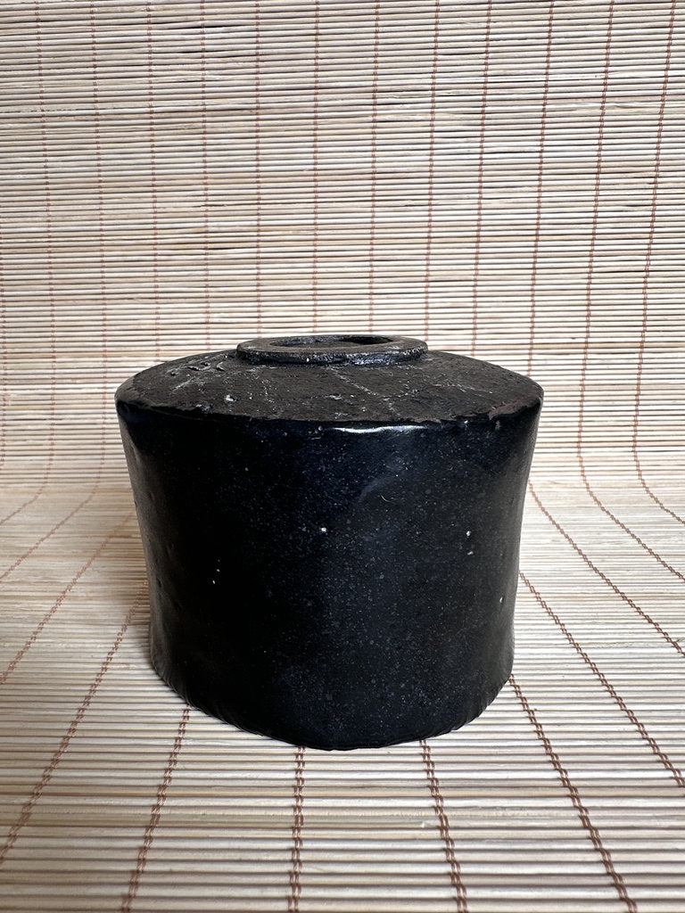A small cylindrical black ceramic pot or vase with a small top opening, placed on a woven straw mat against a matching woven backdrop.