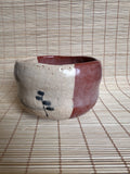 A handmade ceramic bowl with a split beige and reddish-brown glaze, featuring a small black painted motif on the front, set against a bamboo mat background.