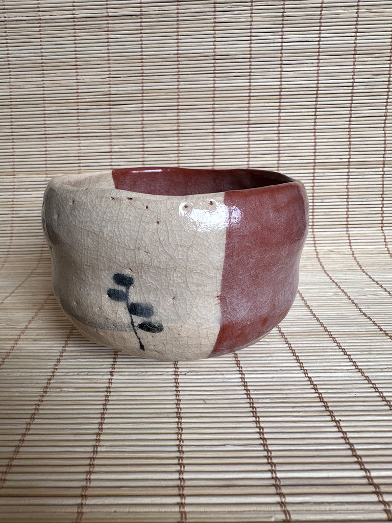 A handmade ceramic bowl with a split beige and reddish-brown glaze, featuring a small black painted motif on the front, set against a bamboo mat background.