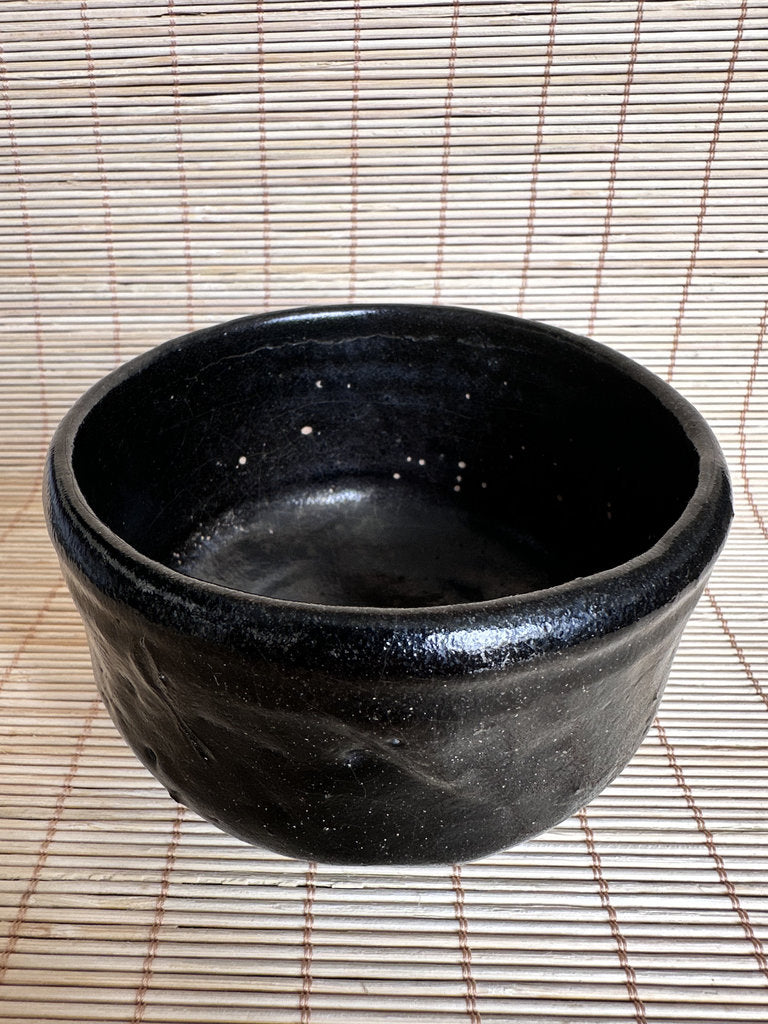 A handmade black ceramic bowl with a textured, hand-painted finish and small white speckles, shown on a woven straw mat.