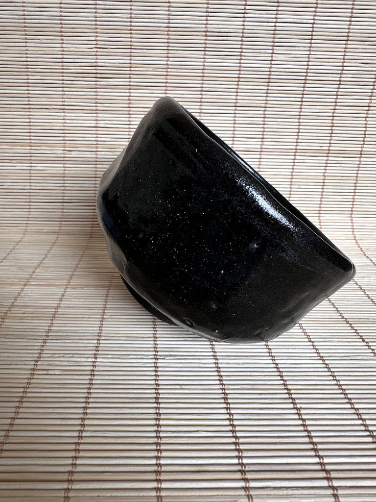 A glossy black ceramic bowl with a speckled glaze, photographed on a beige woven mat background.