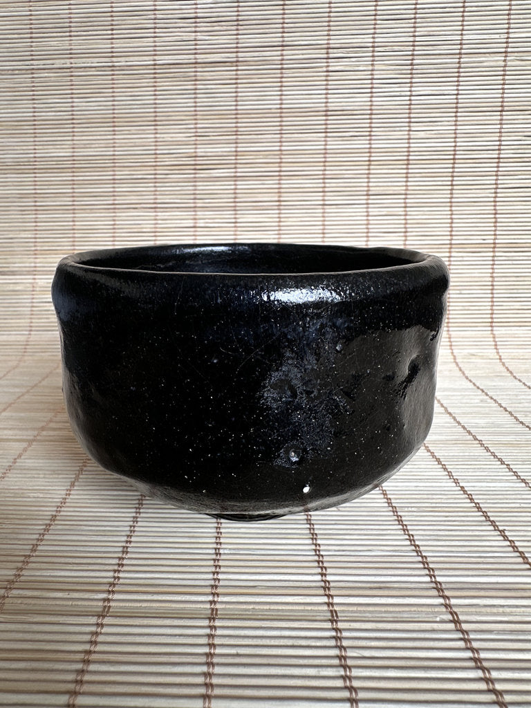 A handmade black ceramic bowl with a glossy, speckled glaze, resting on a light brown bamboo mat.