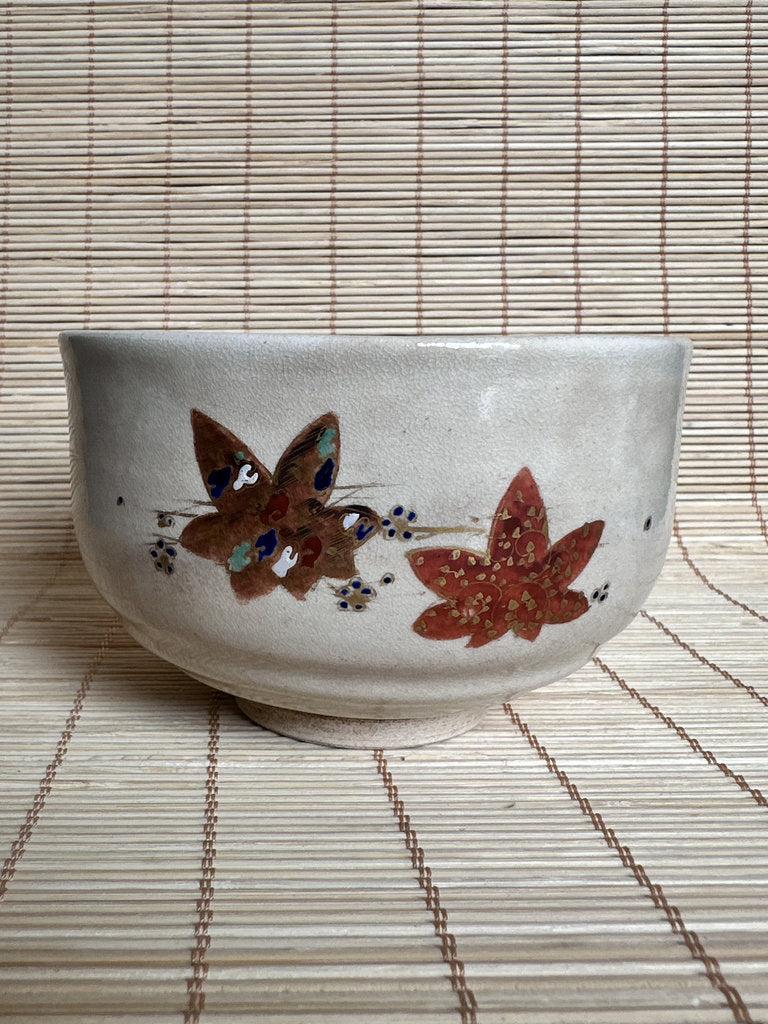 A handmade ceramic bowl with a textured off-white glaze, decorated with a brown butterfly and a red maple leaf motif, resting on a woven straw mat.