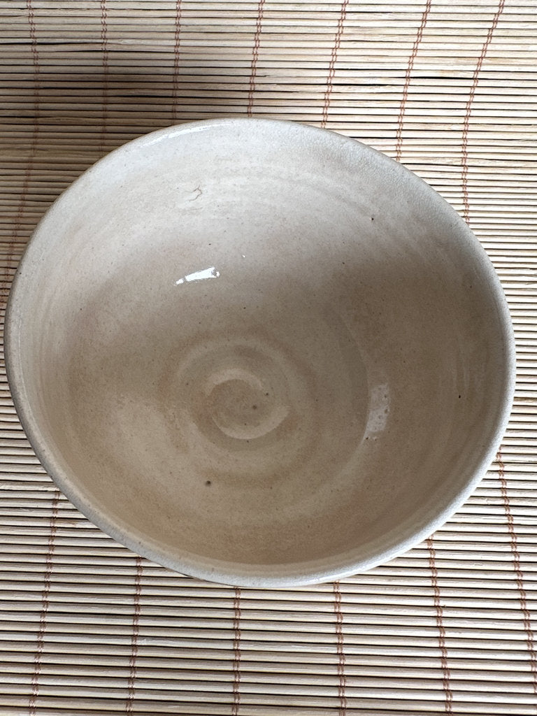 A handmade ceramic bowl with a light beige, matte glaze, photographed from directly above on a bamboo mat.