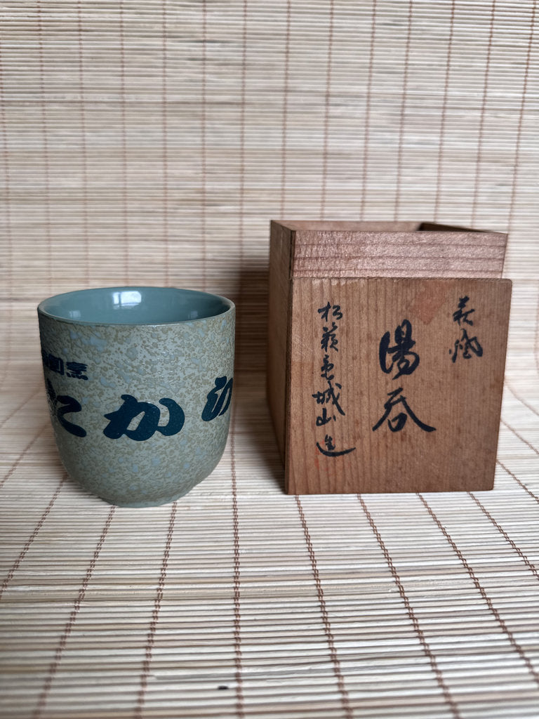 A small speckled ceramic cup with a pale blue interior and dark blue Japanese calligraphy, placed beside a rectangular wooden box with black calligraphy on a woven straw mat background.
