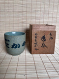 A small speckled ceramic cup with a pale blue interior and dark blue Japanese calligraphy, placed beside a rectangular wooden box with black calligraphy on a woven straw mat background.