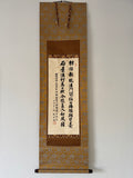 Traditional calligraphy scroll on a decorative gold and brown frame against a white wall.