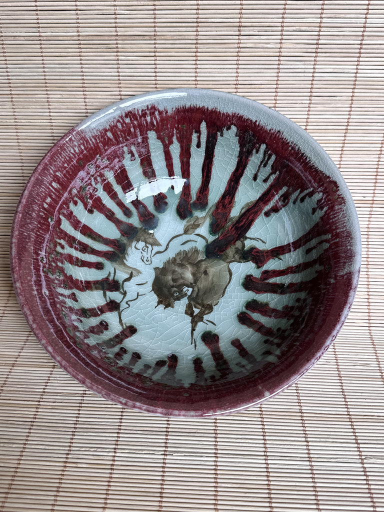 A handmade ceramic bowl with a glossy glaze, featuring a deep reddish-brown painted radial pattern and a central floral motif, resting on a light brown bamboo mat.