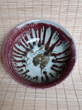A handmade ceramic bowl with a glossy glaze, featuring a deep reddish-brown painted radial pattern and a central floral motif, resting on a light brown bamboo mat.