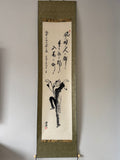 Traditional Japanes ink painting on a vertical scroll with calligraphy against a plain wall.