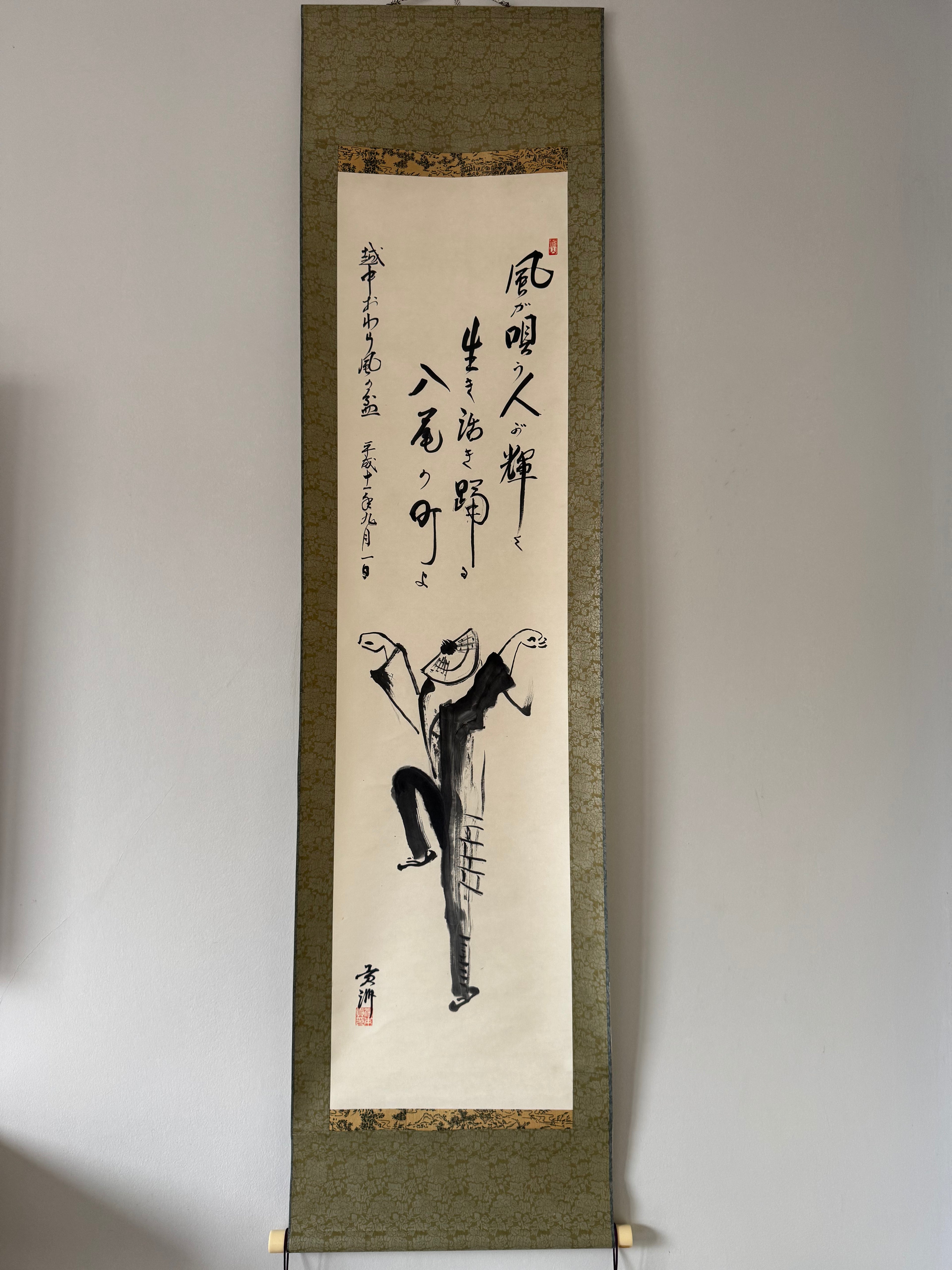 Traditional Japanes ink painting on a vertical scroll with calligraphy against a plain wall.