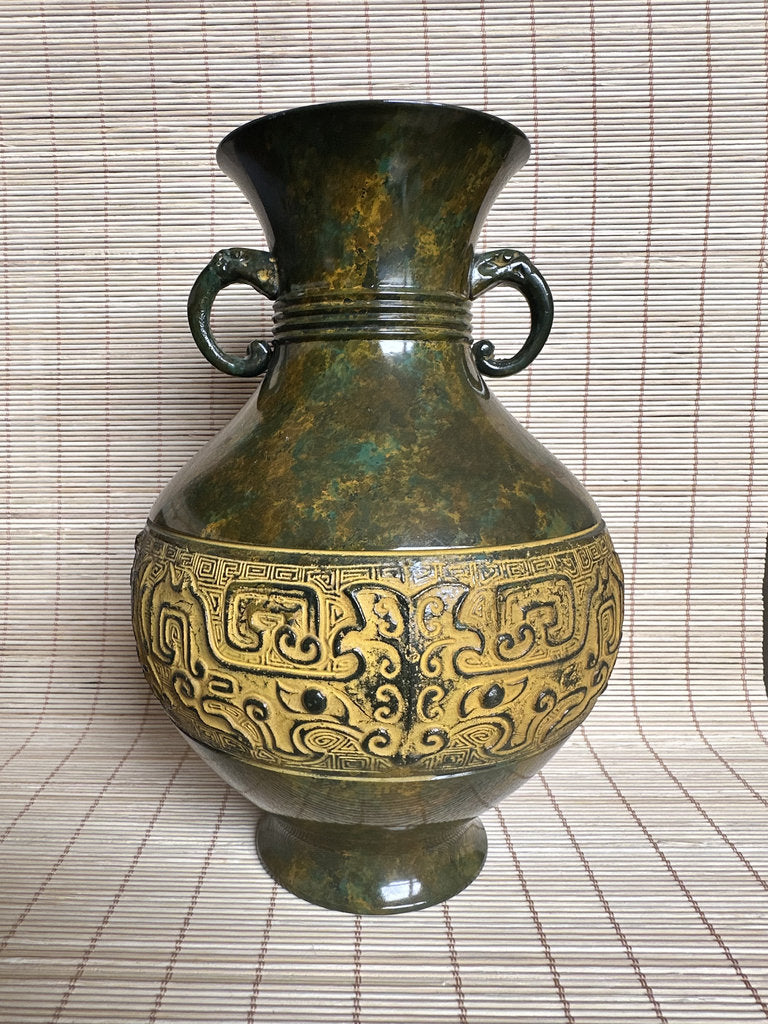 An antique-style bronze vase with a dark green patina and gold-colored Chinese dragon motifs, featuring two side handles, set against a beige woven mat background.
