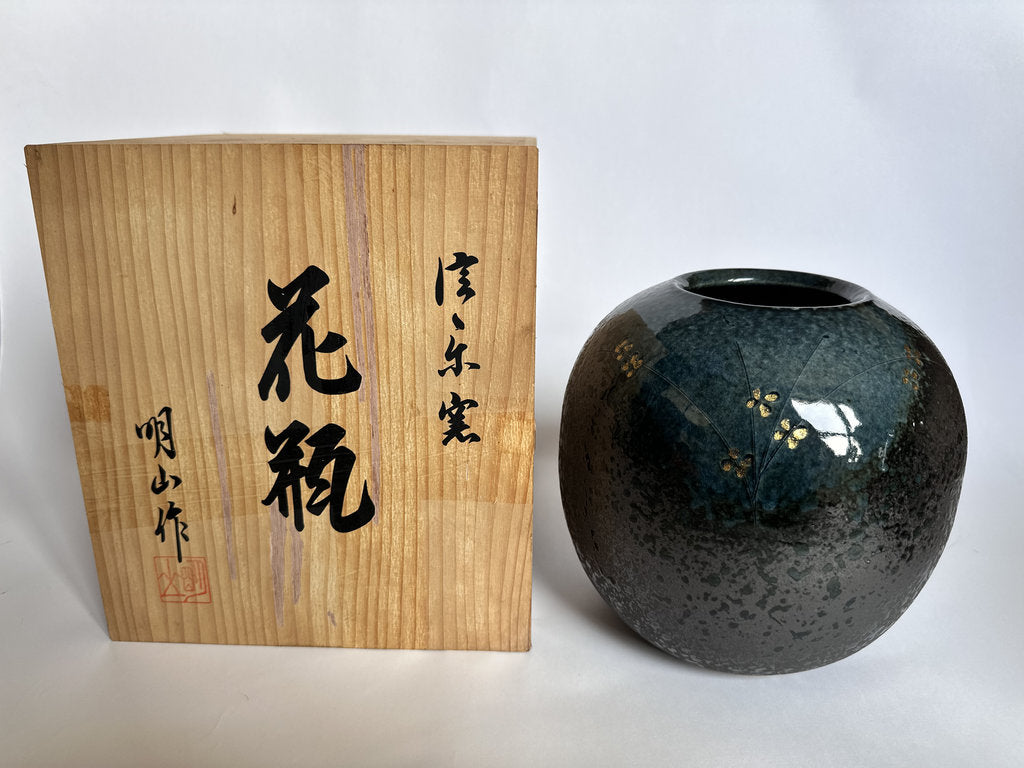 A dark glazed ceramic vase with gold floral details stands next to its light wood presentation box with black calligraphy, photographed against a plain white background.